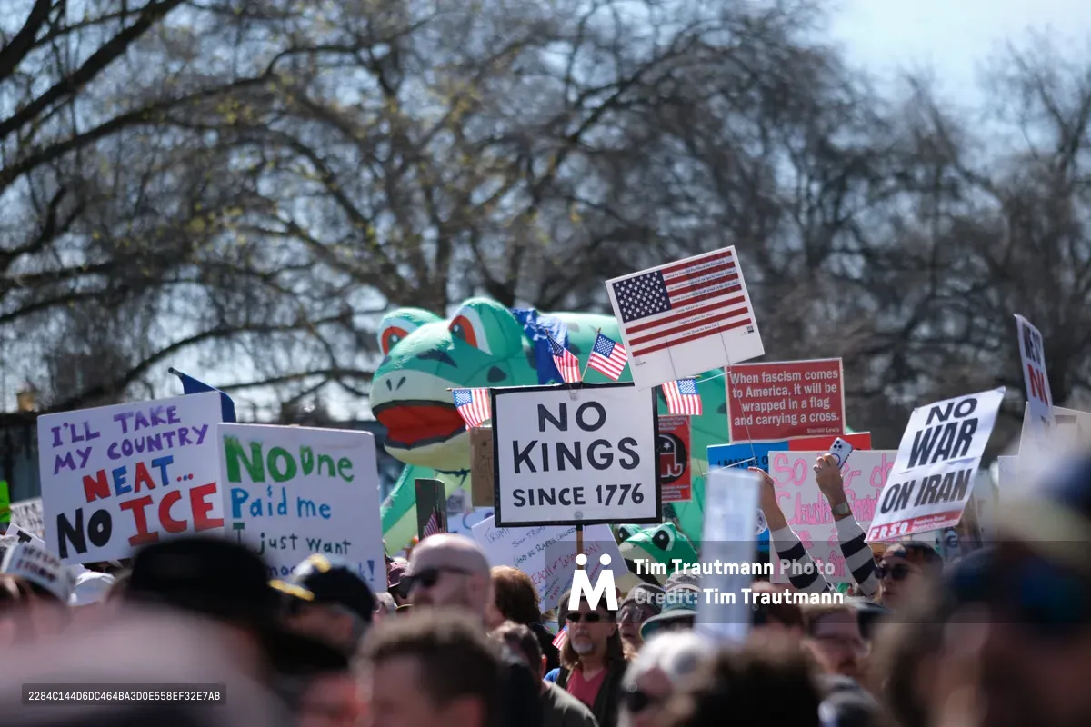 A sea of protesters fills Southwest Ankeny Street in Portland's Old Town district, their handmade signs creating a tapestry of civic dissent against the skeletal winter branches overhead. The crowd brandishes messages like "NO KINGS SINCE 1776" and "I'LL TAKE MY COUNTRY NEAT... NO ICE" alongside American flags and colorful inflatable props, while the soft overcast light diffuses across the dense gathering. The historic Chinatown backdrop frames this moment of political expression, where individual voices merge into a collective chorus of democratic ideals.