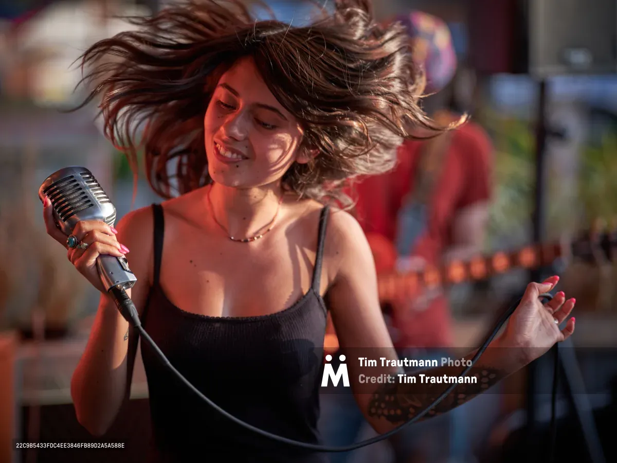 Singer Aubrey Gordon commands the stage with passionate intensity at Portland Mercado in Southeast Portland, her flowing brunette hair caught mid-motion as she grips a vintage-style microphone. Warm amber stage lighting bathes her performance in golden tones, creating dramatic shadows that emphasize the raw energy of her live set. The intimate venue setting frames her dynamic presence as she delivers her performance with closed eyes and expressive gestures, fully immersed in the music.