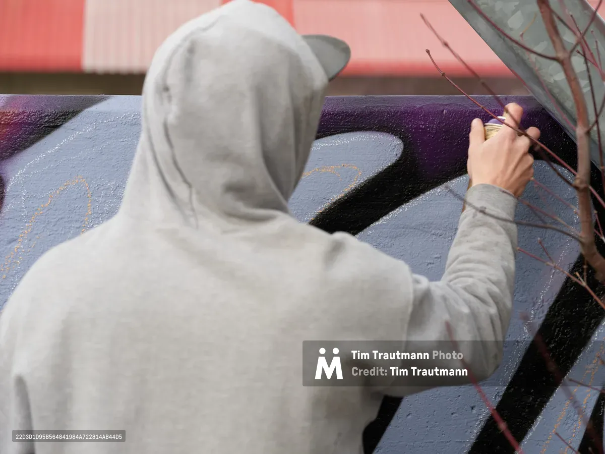Travis Fields, known as Campographic, works intently on a vibrant mural at the Clay Creative Building in Portland's Hosford-Abernethy neighborhood. Dressed in a light gray hoodie, the artist's focused gestures guide crimson details across bold purple and blue geometric forms. The afternoon light catches the textured spray paint surface as creative energy transforms the urban canvas.