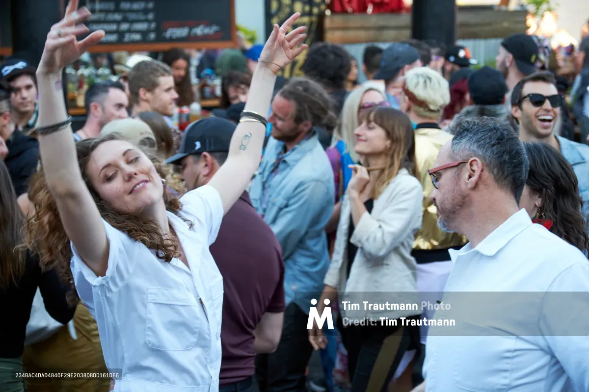 A young woman with curly brown hair raises her arms in pure joy, eyes closed in musical bliss as she dances among a packed crowd at White Owl Social Club. The intimate Portland venue pulses with afternoon energy as dozens of revelers move to the rhythm, their faces illuminated by warm natural light filtering through the space. The scene captures the uninhibited spirit of a summer day party, where strangers become temporary dance partners in the communal celebration of music.