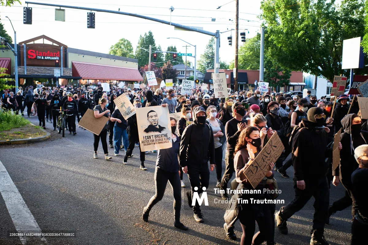 Hundreds of demonstrators flood Northeast 33rd Avenue in Portland's Laurelhurst neighborhood during a Black Lives Matter protest following George Floyd's murder. Masked protesters carry handmade cardboard signs bearing messages like "I Can't Breathe" and "Black Lives Matter" as they march past the Standard restaurant under overcast evening skies. The diverse crowd moves peacefully through the tree-lined commercial district, their collective presence transforming the quiet neighborhood street into a powerful corridor of social justice advocacy.