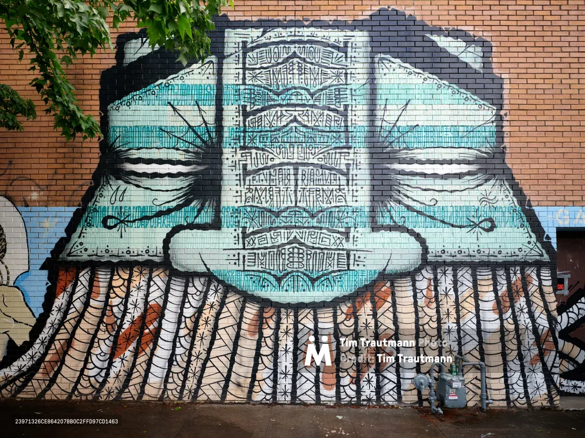 A striking abstract composition dominates this street mural on Southeast Division Street in Portland's Richmond neighborhood, where the historic Oregon Theater once stood. The artwork features flowing forms rendered in turquoise and earth tones against warm brick, incorporating geometric patterns and organic shapes. Dappled sunlight filters through overhanging tree branches, casting organic shadows across the carefully detailed abstract design that transforms the urban streetscape into a canvas of artistic expression and neighborhood identity.