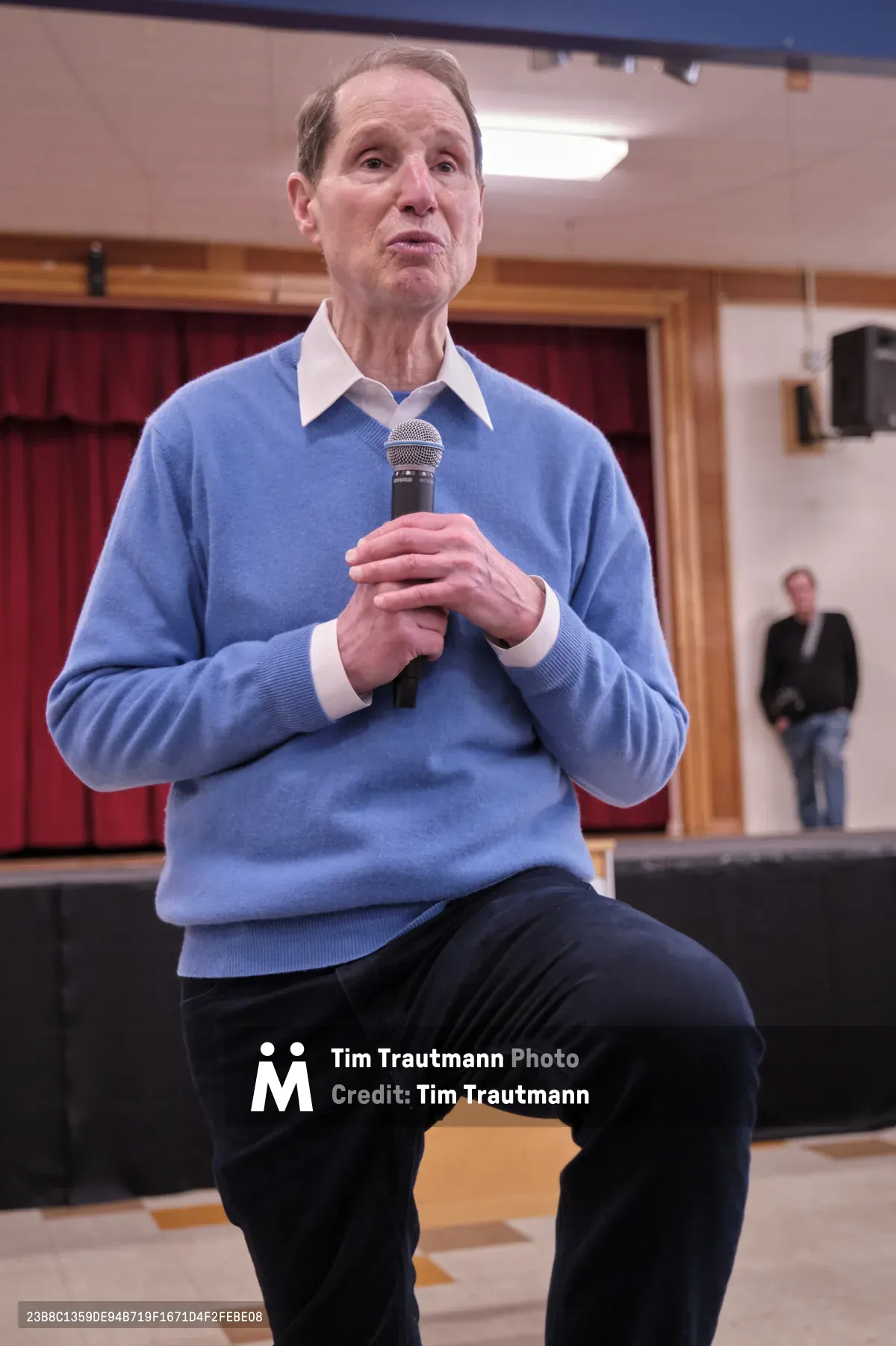 Senator Ron Wyden leans forward intently while speaking into a handheld microphone at a town hall meeting in Robert Gray Middle School's gymnasium in Portland, Oregon. The senior politician, dressed in a powder blue sweater over a crisp white collared shirt, commands attention from his seated position on the gymnasium's bleachers. Fluorescent lighting casts an even glow across the institutional setting, where red curtains frame wooden doors in the background and a lone attendee waits in the distance, creating an intimate yet formal atmosphere for civic engagement.