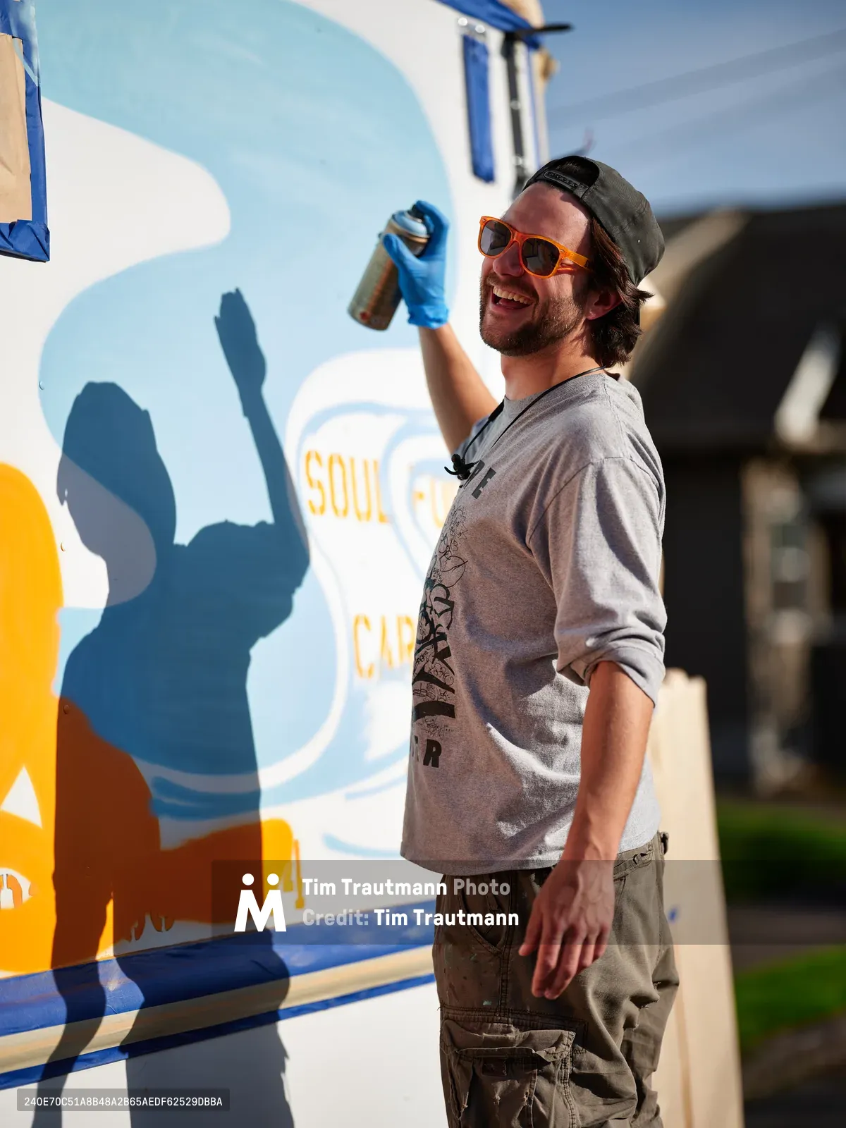 A bearded muralist in orange sunglasses and backwards cap raises a spray paint can in triumph beside his vibrant food truck artwork in Northeast Portland. Golden afternoon light illuminates his genuine smile and the bold blue and orange mural behind him, casting dramatic shadows that amplify the creative energy of this urban art scene. The artist's casual attire and relaxed pose speak to the authentic street art culture thriving in this corner of the Pacific Northwest.