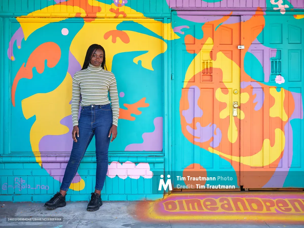 Naomi Likayi stands confidently beside her completed street mural on Northeast 15th Avenue in Portland's Alberta district, her signature visible in the corner of the psychedelic artwork. The young Black artist wears a striped turtleneck and jeans against the explosion of turquoise, orange, yellow and purple abstract forms that transform the building's facade. Warm afternoon light illuminates both the artist and her kaleidoscopic creation, capturing a moment of artistic completion in one of Portland's most vibrant neighborhoods.