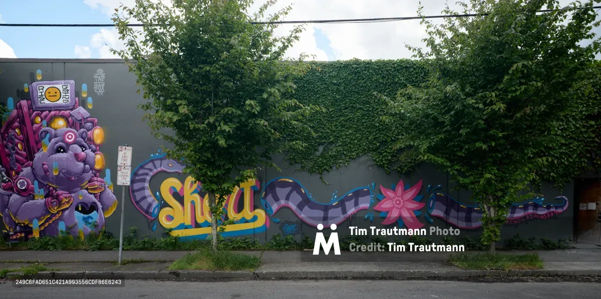 A vibrant street mural dominates a concrete wall along Northeast Irving Street in Portland's Kerns neighborhood, where cascading ivy from overhead trees creates a natural frame. The centerpiece features a whimsical purple robot character surrounded by geometric patterns, tentacle-like forms, and colorful lettering that spells "Shay" in bold yellow script. The artwork blends cyberpunk aesthetics with playful cartoon elements, creating a striking contrast against the gray urban landscape beneath scattered clouds.