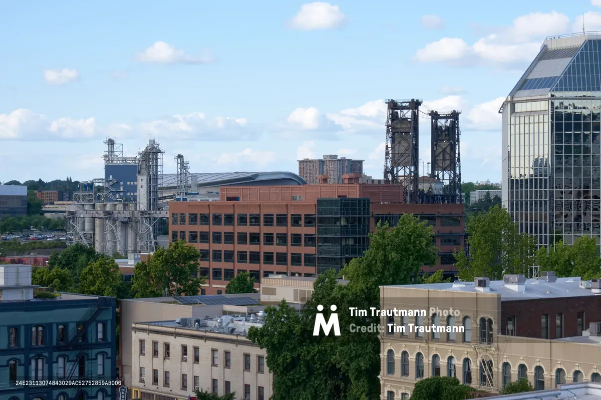 An elevated view across Portland's historic Chinatown district reveals a compelling juxtaposition of architectural eras under a partly cloudy afternoon sky. The foreground showcases traditional brick and masonry buildings with arched windows, while mid-ground industrial structures including towering grain silos and processing facilities create dramatic vertical elements. Modern glass-clad high-rises pierce the horizon, their reflective surfaces catching the natural daylight that bathes this diverse urban landscape in warm, even tones.