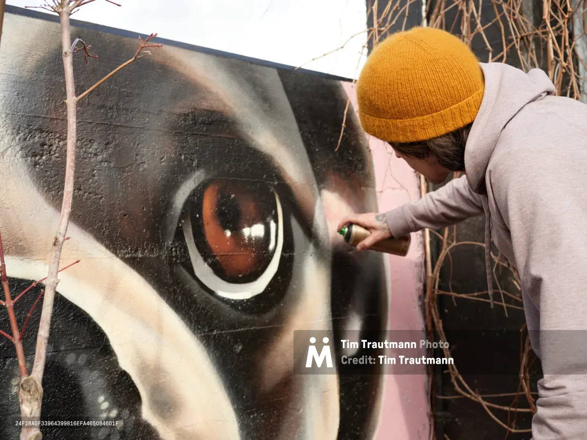 Muralist Wetiko meticulously applies spray paint to a striking hyperrealistic eye mural on an exterior wall in Portland's Hosford-Abernethy neighborhood. Wearing a golden yellow beanie and pale hoodie, the artist works with focused precision on the luminous amber iris, surrounded by detailed eyelashes and photorealistic skin tones. The intimate moment captures the transformation of urban space through art, with dried vegetation creating a natural frame around the creative process.