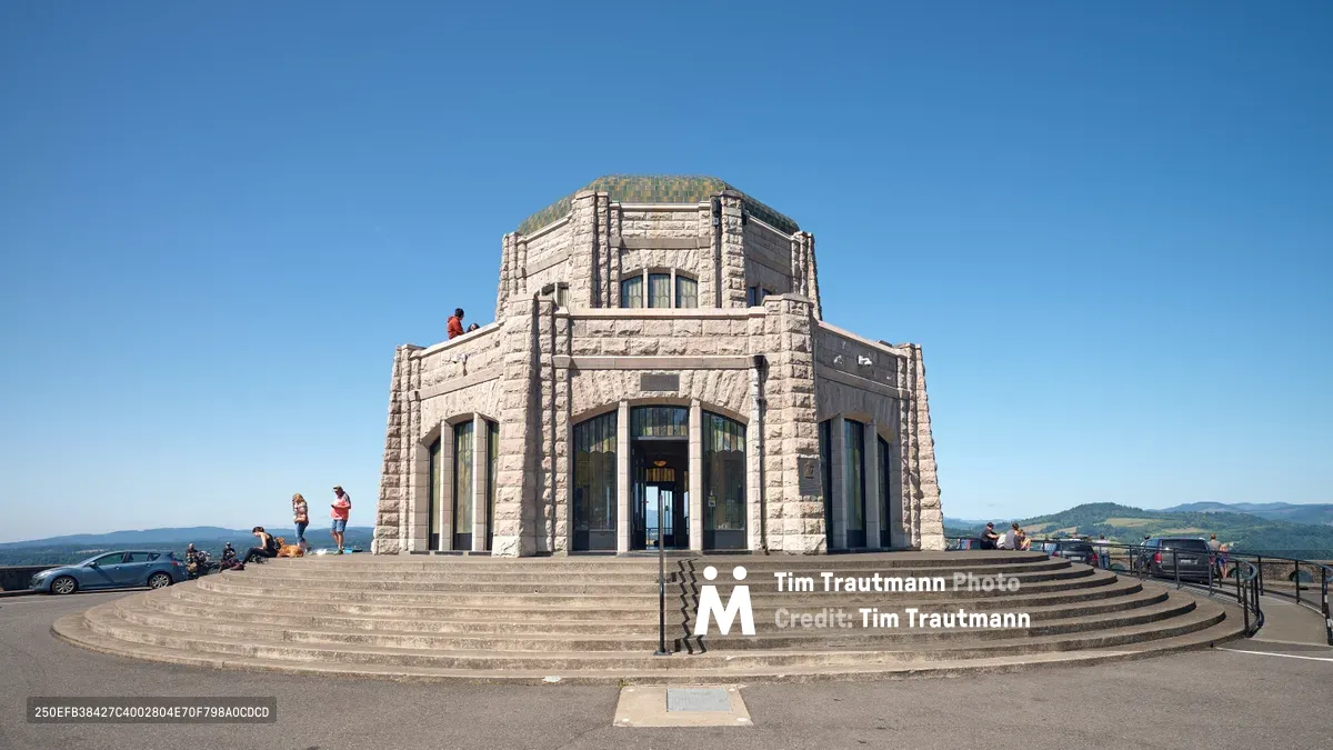 The iconic Vista House stands majestically atop Crown Point, its weathered stone facade gleaming under brilliant blue skies. Visitors gather on the circular stone steps and observation deck, drawn by the panoramic views of the Columbia River Gorge stretching toward distant forested hills. The Art Nouveau monument's distinctive octagonal architecture creates a striking silhouette against the endless Oregon sky, while tourists capture memories and explore this beloved Pacific Northwest landmark.