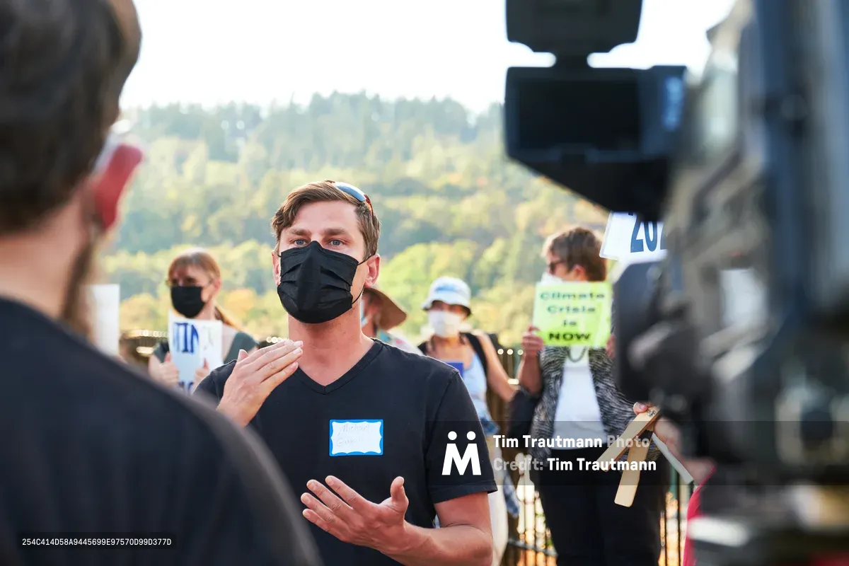 Michael Gaskill, wearing a black face mask and v-neck shirt with a blue name tag, speaks earnestly to a KOIN Channel 6 news crew during a climate change protest at Representative Kurt Schrader's Oregon City office. The interview unfolds against a backdrop of forested hills bathed in golden afternoon light, while fellow demonstrators holding signs gather in the soft-focused background. The composition captures the intersection of grassroots activism and media coverage in the tree-lined McLoughlin Promenade setting.