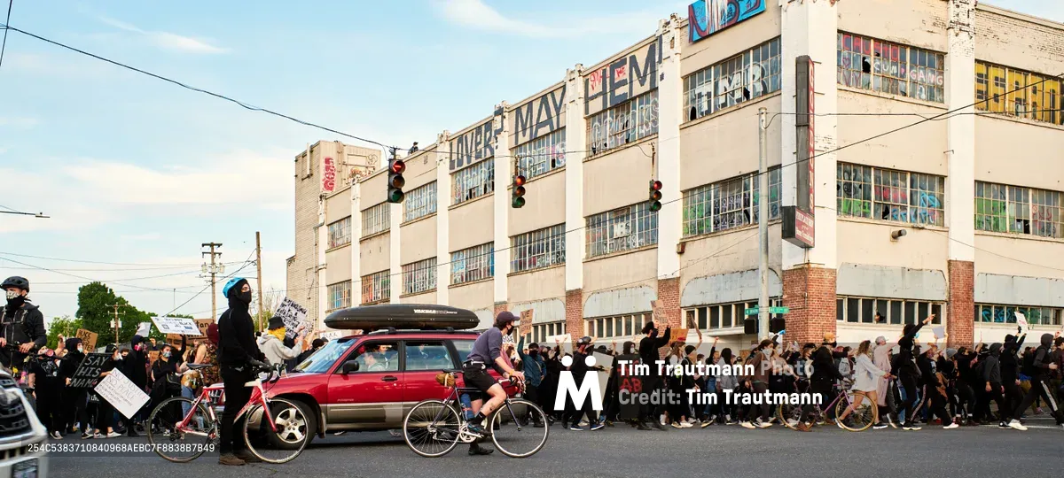 Hundreds of Black Lives Matter protesters flow down Northeast 33rd Avenue in Portland's Grant Park neighborhood, creating a river of activism against the backdrop of weathered industrial buildings. The late afternoon light illuminates handmade signs and the determined faces of demonstrators walking alongside cyclists and a red SUV with a roof cargo box. Power lines crisscross the pale sky above the procession, while the historic May-Em building anchors the urban landscape with its faded signage and multi-story windows.