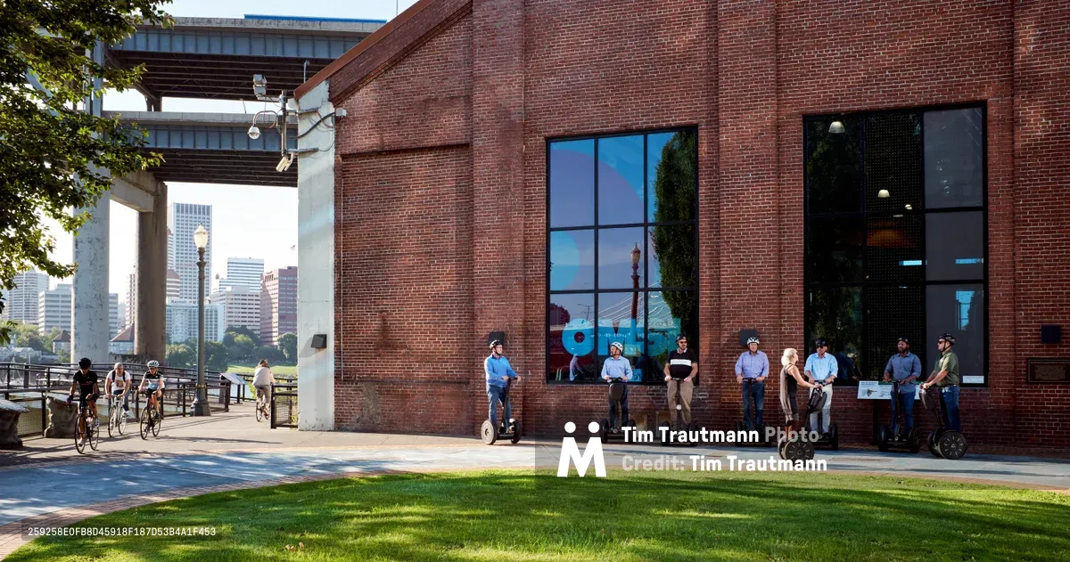 A diverse group of tourists on Segways pauses along the esplanade outside the Oregon Museum of Science and Industry, their helmeted silhouettes arranged against the warm red brick facade. The late afternoon light illuminates the industrial architecture while Portland's downtown skyline rises in the background, creating a perfect juxtaposition of recreational technology and urban exploration. The manicured lawn and concrete walkway frame the scene, capturing a moment of pause in this modern city adventure.