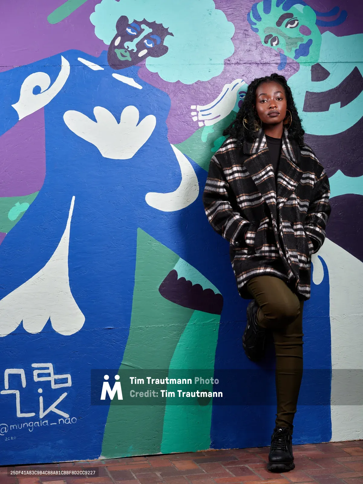 Artist Naomi Likayi stands confidently against her explosive mural at Portland's World Trade Center, wearing a black and white plaid flannel over dark jeans and boots. The artwork behind her bursts with cobalt blues, seafoam greens, and violet purples, featuring abstract faces and flowing forms that create a dynamic backdrop. Her relaxed pose and direct gaze embody the creative spirit that transformed this boarded urban space into a canvas of expression through the Portland Street Art Alliance commission.