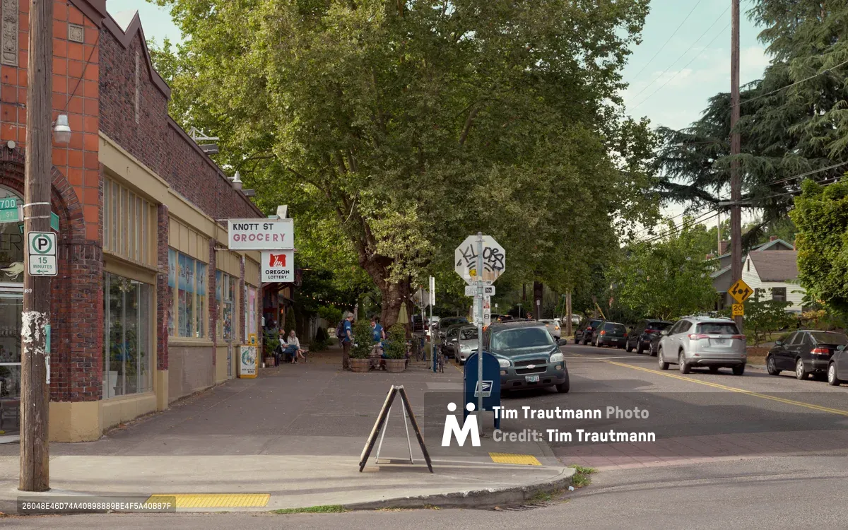 A quiet afternoon captures the essence of Northeast Knott Street in Portland's Irvington neighborhood, where red brick storefronts house local businesses like Knott Street Grocery. Mature trees create a canopy over the two-lane street as residents gather on sidewalks and cars navigate the tree-lined thoroughfare. The soft, diffused light suggests late afternoon, casting gentle shadows across the pedestrian-friendly streetscape that exemplifies Portland's walkable neighborhood character.