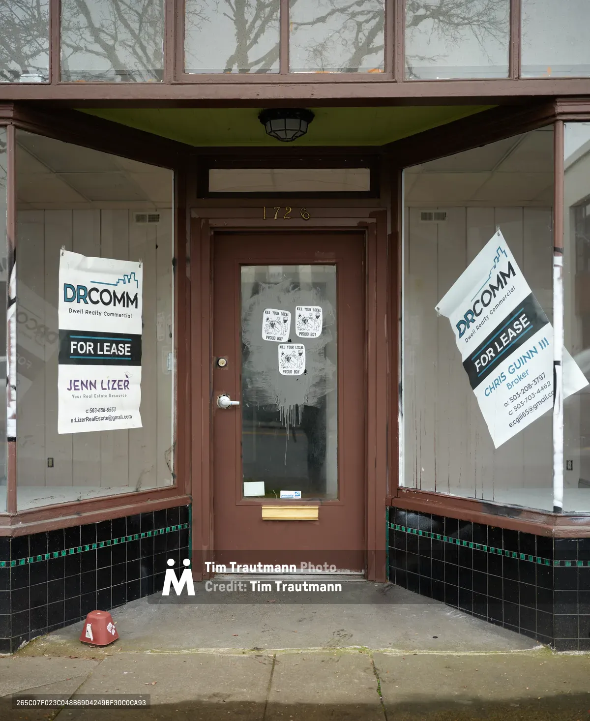 A weathered storefront at 1726 Northeast Alberta Street displays the changing face of Portland's historically Black Alberta District. Multiple real estate signs from DRCOMM and agent Jenn Lizer advertise commercial space for lease, while mysterious posters blur behind the glass door of what was once Earl's Barber Shop. The building's modest architecture - brown wood trim, green ceiling accents, and black tile wainscoting - speaks to the neighborhood's working-class heritage now caught between preservation and development.
