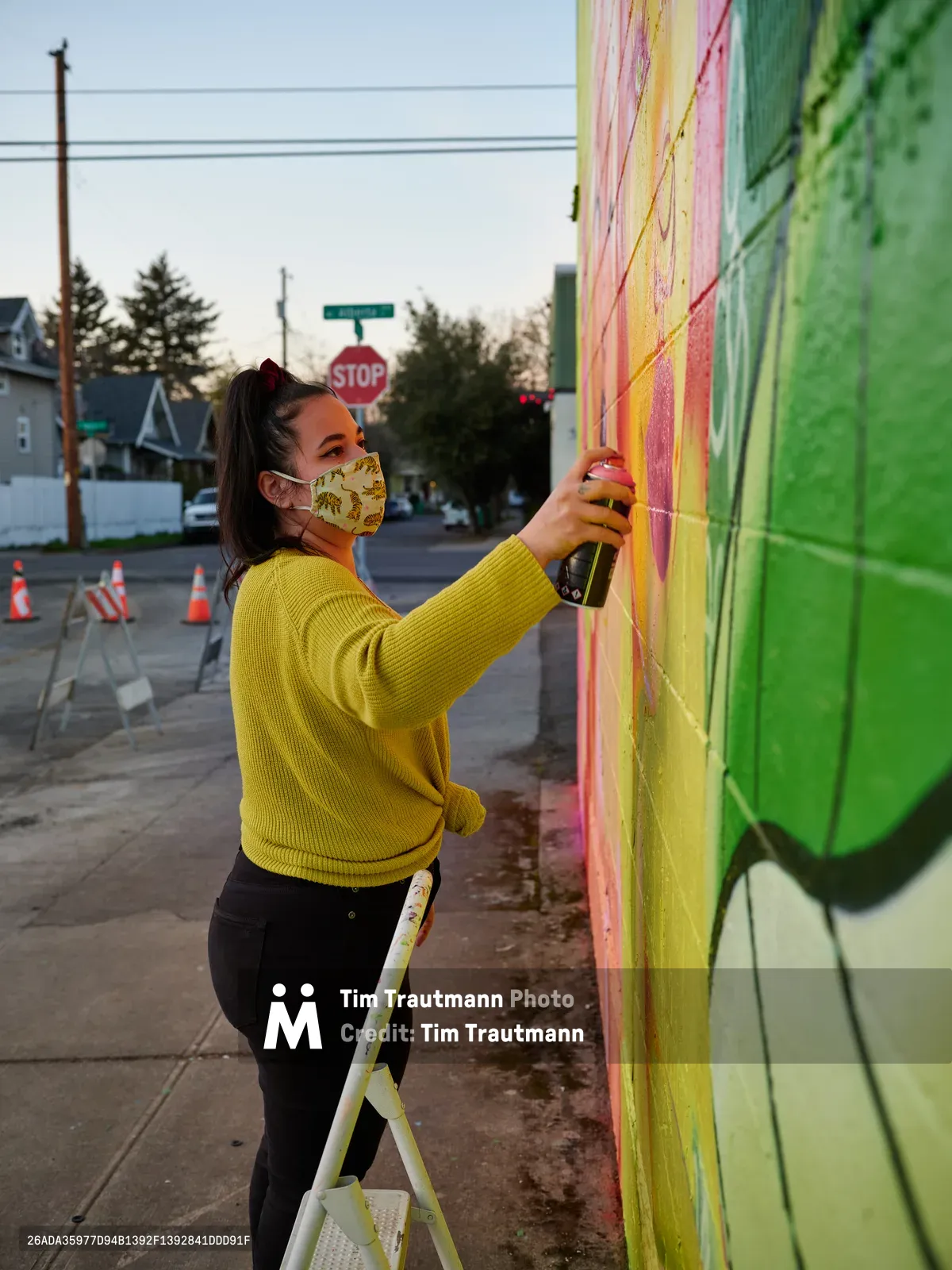 Multidisciplinary artist Villatose applies spray paint to her colorful Jardín Cubana mural featuring three female figures along Northeast Alberta Street in Portland's Alberta Arts District during golden hour. Wearing a bright yellow sweater and floral face mask, she works methodically from a small ladder, adding layers of pink, yellow, and green paint to transform the urban wall. The quiet residential street stretches behind her, punctuated by traffic cones and power lines against the soft evening sky.