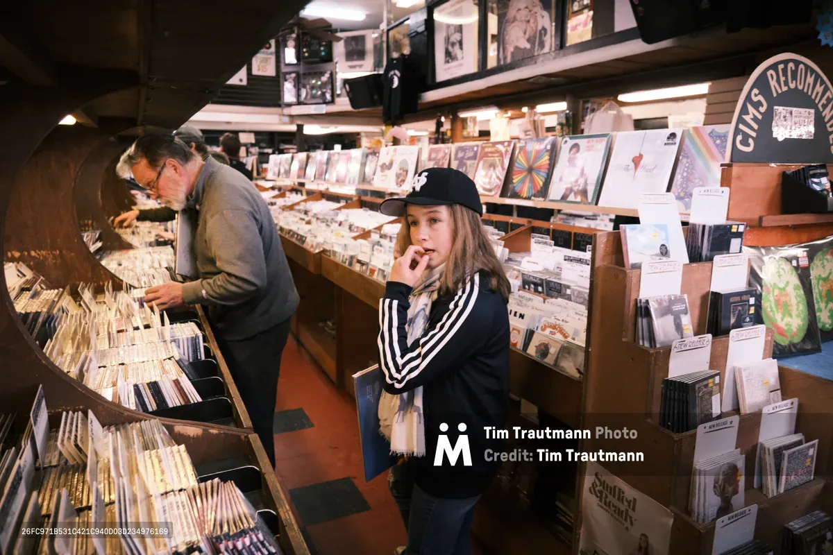 Inside the legendary Music Millennium record shop on East Burnside Street in Portland's Kerns neighborhood during Record Store Day, two generations of music lovers browse through carefully curated vinyl collections. A middle-aged man in glasses and gray sweater methodically flips through albums in wooden crates, while a young woman in a black baseball cap and striped jacket contemplates her next discovery. Warm fluorescent lighting illuminates towering shelves packed with records, creating an intimate atmosphere of musical exploration as fans hunt for limited Record Store Day releases in this iconic Oregon institution.