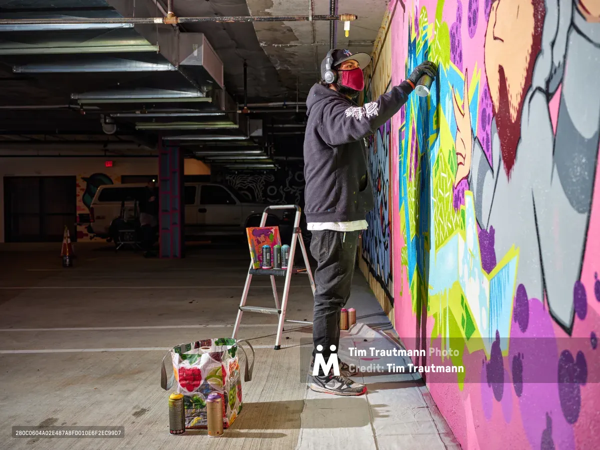 In the industrial depths of Clay Creative's Southeast Portland parking garage, a masked muralist applies brilliant strokes of magenta, lime green, and azure to transform concrete walls into living art. The artist, wearing headphones and a crimson face covering, stands atop a metal stepladder surrounded by scattered spray cans and paint containers. Exposed ductwork and fluorescent lighting cast harsh shadows across the cavernous space, while the emerging mural bursts with color against the stark urban infrastructure.