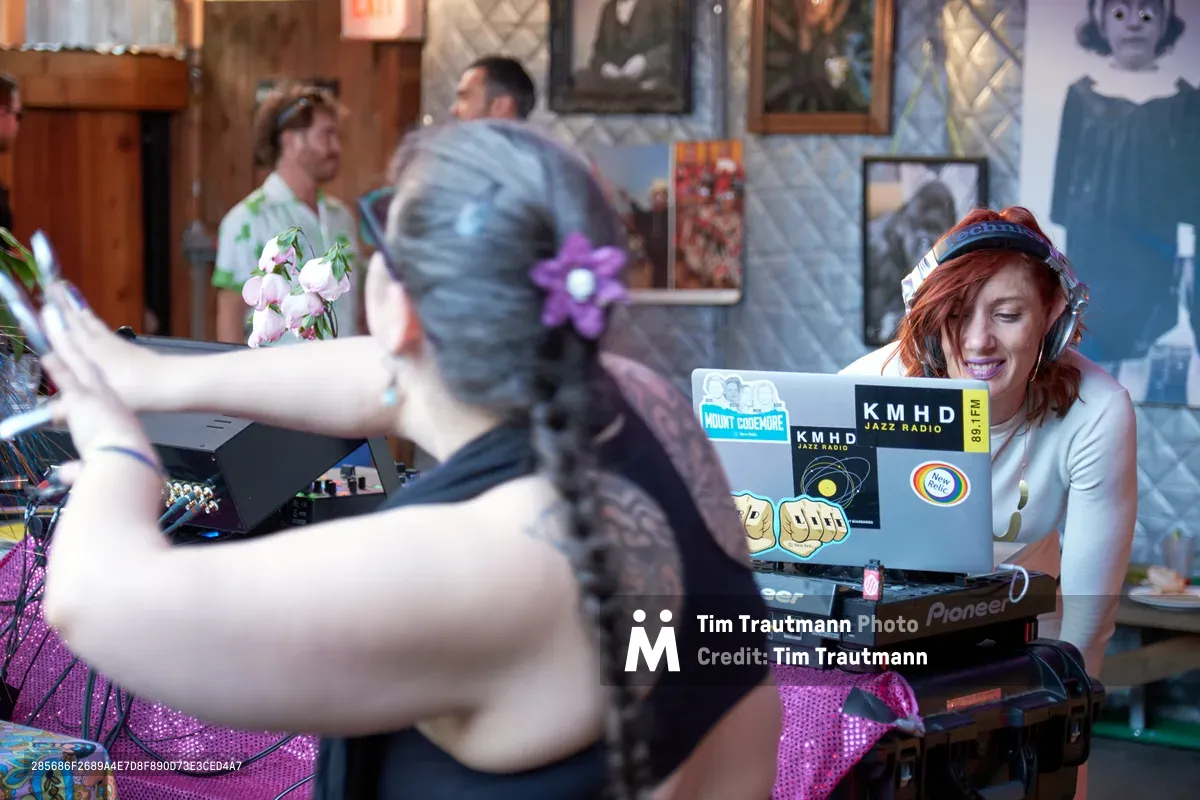 DJ Doctor Dakar commands the decks at White Owl Social Club's Your Sunday Best season opener, her auburn hair catching the ambient light as she focuses intently on her Pioneer setup. The intimate Portland venue pulses with energy as partygoers dance in the background, their movement creating dynamic blur against the eclectic wall art and quilted booth seating. Stickers from KMHD Jazz Radio and other music collectives decorate her laptop, testament to the underground music scene's collaborative spirit.