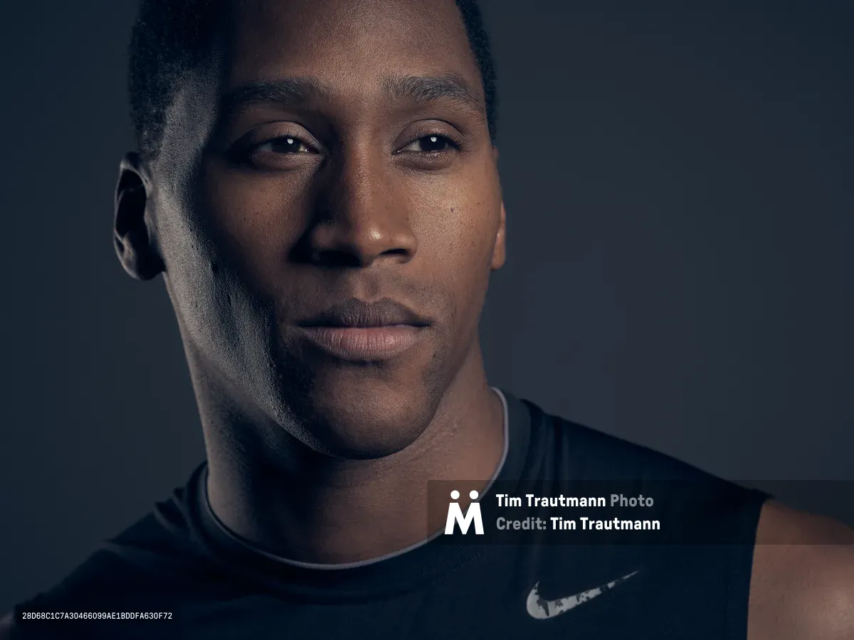 A close-up studio portrait of a young man wearing a black Nike sleeveless shirt, lit dramatically against a dark grey background. He gazes slightly off-camera with a calm, composed expression.