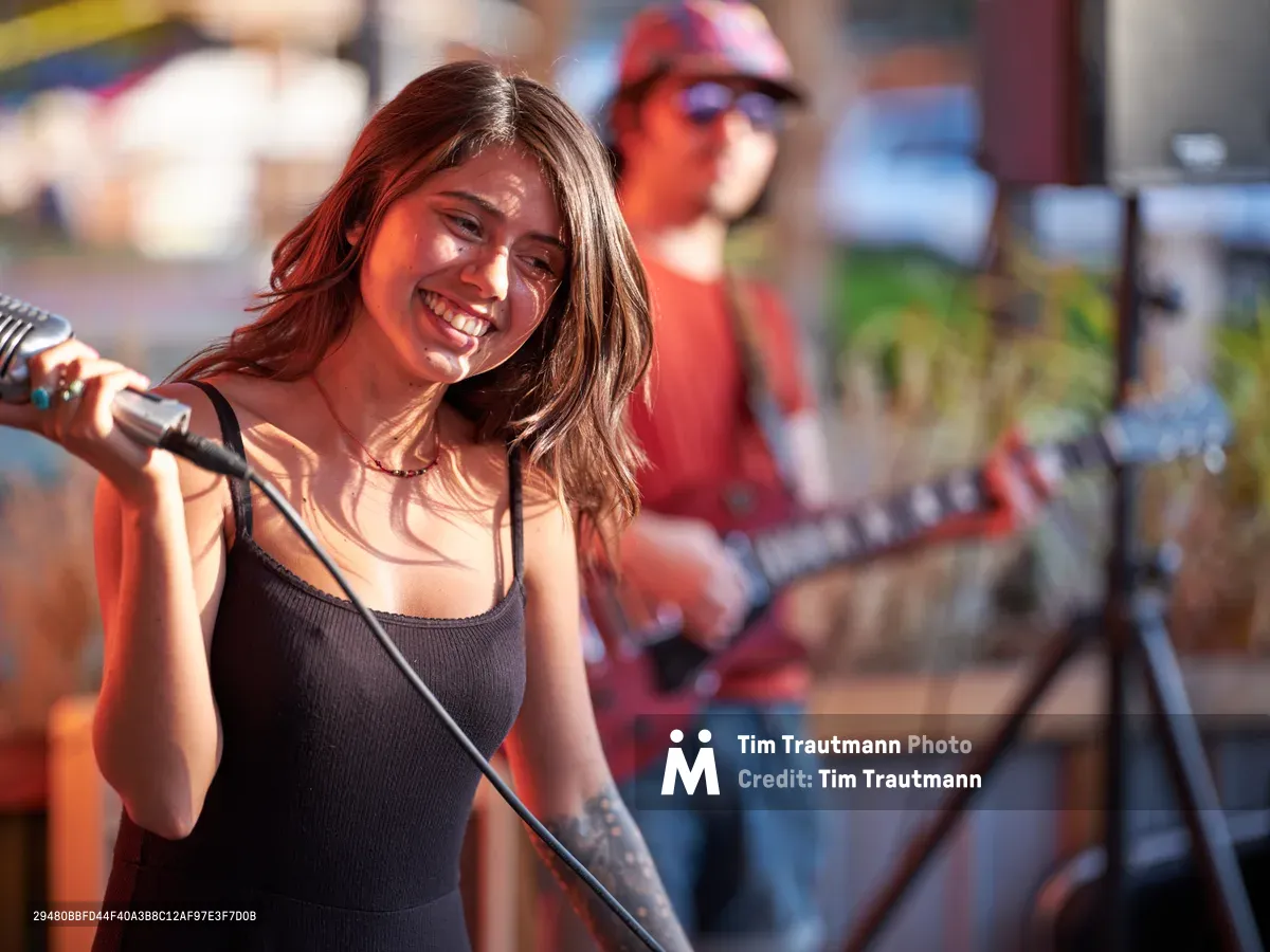 Aubrey Gordon radiates joy as she performs at Portland Mercado in Southeast Portland, her tattooed arm gripping the microphone while golden afternoon light bathes her face. The young vocalist wears a sleek black tank top, her wavy brown hair catching the warm sunlight as she delivers her set to the gathered crowd. Behind her, a guitarist in a red cap provides accompaniment, creating an intimate outdoor performance atmosphere in this vibrant multicultural food market.