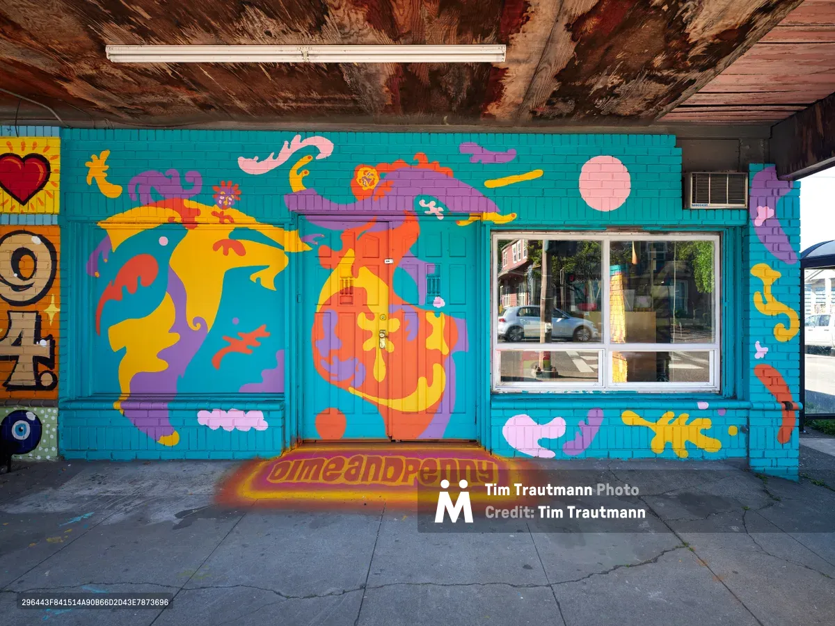 A vibrant street mural by artist Naomi Likayi transforms a weathered storefront wall in Portland's Alberta neighborhood. The turquoise backdrop hosts a fantastical composition of abstract human figures, floating islands, and whimsical characters rendered in bold oranges, purples, and yellows. Exposed wooden ceiling beams frame the artwork above, while cracked concrete stretches below, creating a compelling contrast between urban decay and artistic renewal.