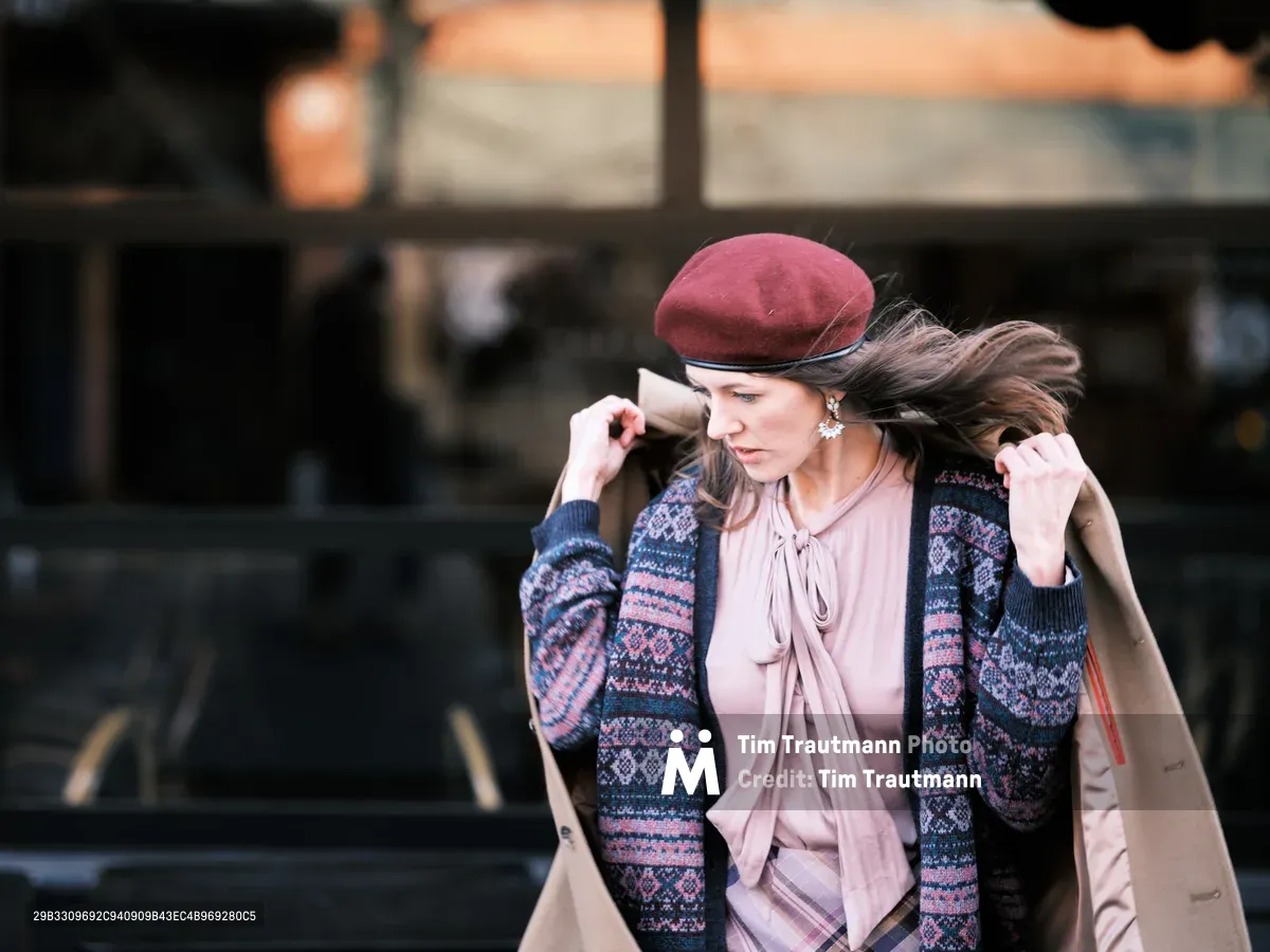 A young woman adjusts her burgundy beret against the wind while walking through Portland's Alberta Arts District. Her layered ensemble of a Fair Isle cardigan over a blush peasant blouse creates textural contrast against the industrial backdrop of Wells Glass Studio. The candid moment captures her in mid-stride, hair flowing with the breeze as she grips her camel coat, embodying the effortless sophistication of urban autumn fashion.