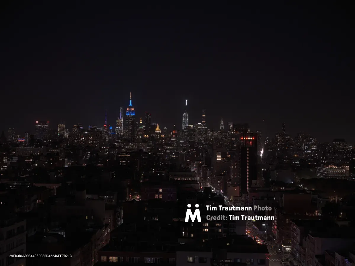 The iconic Manhattan skyline emerges from the darkness, viewed from the elevated perspective of Chinatown's Canal Street corridor. The Empire State Building stands prominently illuminated in blue against the deep twilight sky, while scattered windows across countless residential and commercial buildings create a constellation of warm light. A busy street cuts through the foreground, its traffic creating streaks of red and white light that guide the eye toward the towering skyscrapers of Midtown Manhattan.