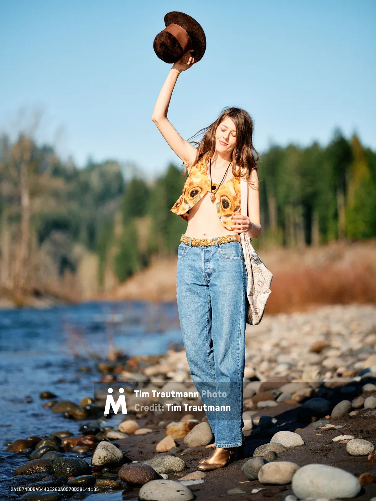 A young woman in vintage-inspired attire raises her wide-brimmed hat triumphantly along the rocky shoreline of Sandy River in Oregon's Floodplain Trail. The golden hour light illuminates her sunflower-print vest and creates a warm, nostalgic atmosphere as gentle waters lap against the smooth river stones. Her joyful gesture and closed eyes convey a moment of pure liberation and connection with nature.