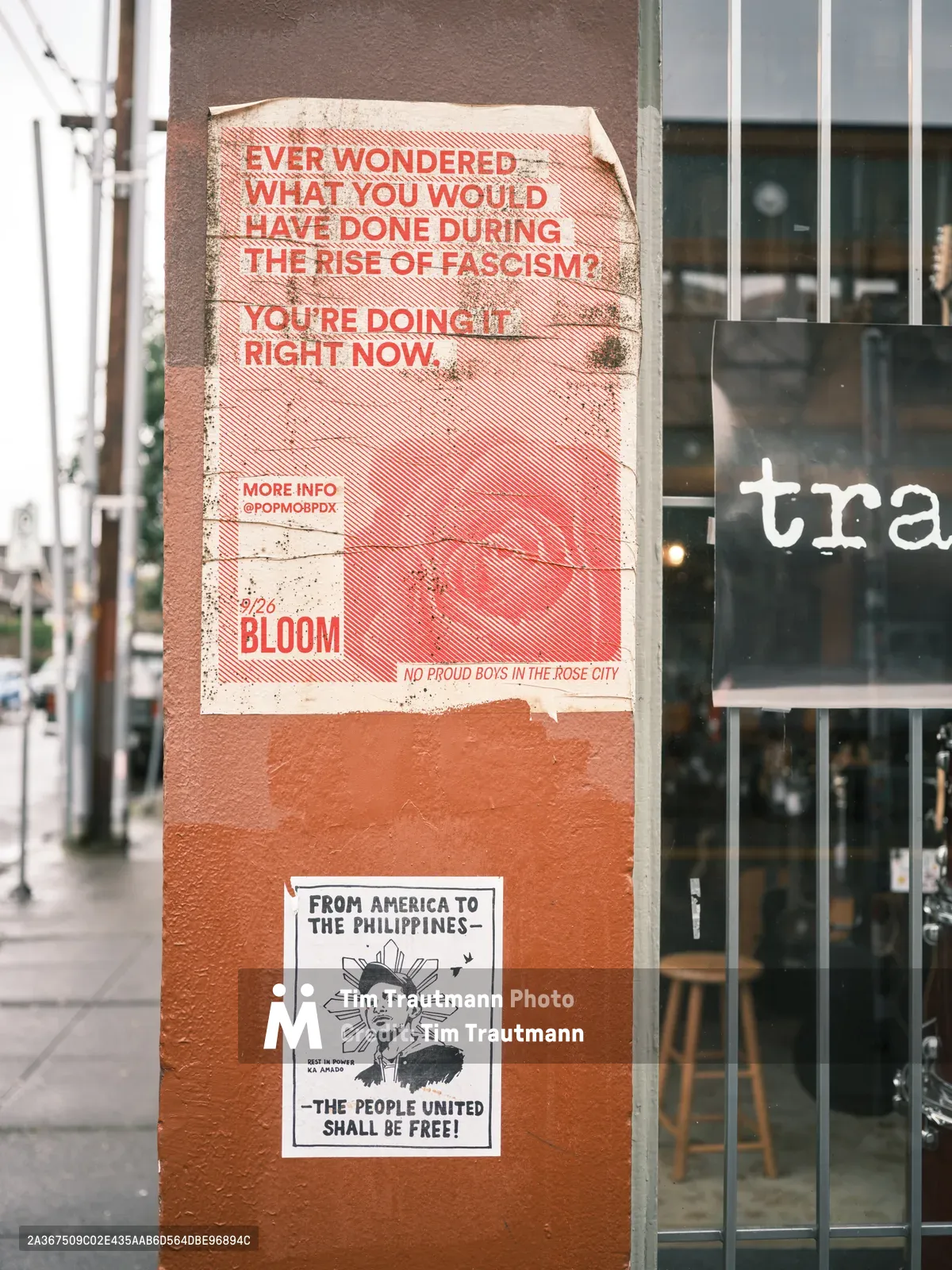 Two provocative political posters adhere to a weathered utility pole on Northeast Alberta Street in Portland's Alberta Arts District. The upper poster, printed in bold red and white graphics, poses a stark question about fascism and complicity, while below it, a black and white flyer featuring imagery reminiscent of the Statue of Liberty connects American and Filipino liberation movements. The compositions are captured against the backdrop of storefront windows, creating a layered urban narrative of dissent and activism.