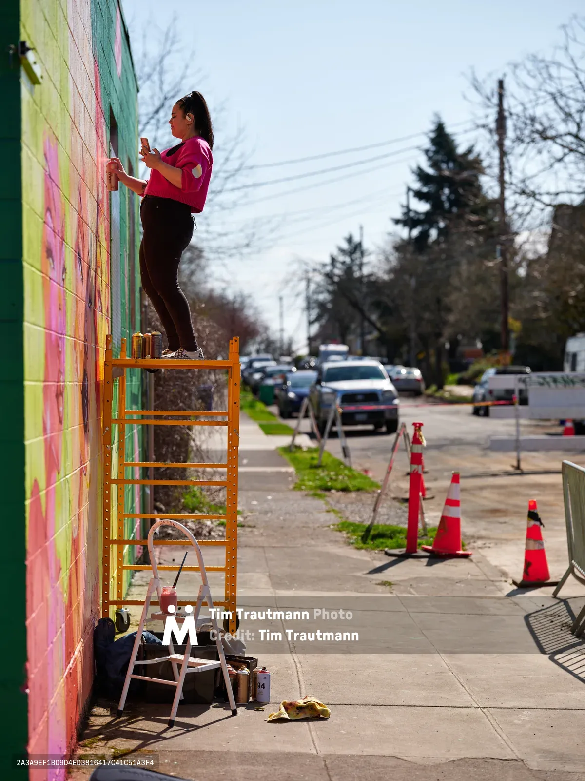 Multidisciplinary artist Vilatose stands atop scaffolding, spray paint in hand, adding bold strokes to a rainbow-hued mural cascading down a brick wall along Northeast Alberta Street. The afternoon light illuminates her focused expression and magenta shirt as she works against the backdrop of Portland's Alberta Arts District, where orange traffic cones mark the creative transformation in progress. Paint supplies and a stepladder scatter the sidewalk below, evidence of the urban canvas taking shape.