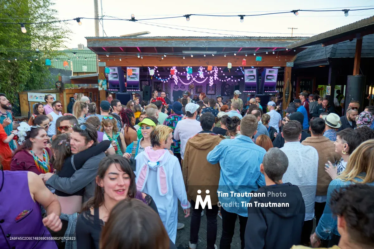 A vibrant crowd fills the outdoor patio of White Owl Social Club in Portland, Oregon, during the season opener of Your Sunday Best, a popular summer day party. Revelers in colorful costumes and casual attire dance beneath string lights and festive decorations, with the venue's covered stage area glowing with purple lighting in the background. The late afternoon atmosphere captures the essence of Portland's eclectic music scene, as dozens of party-goers embrace the celebratory mood of the season's first outdoor dance gathering.