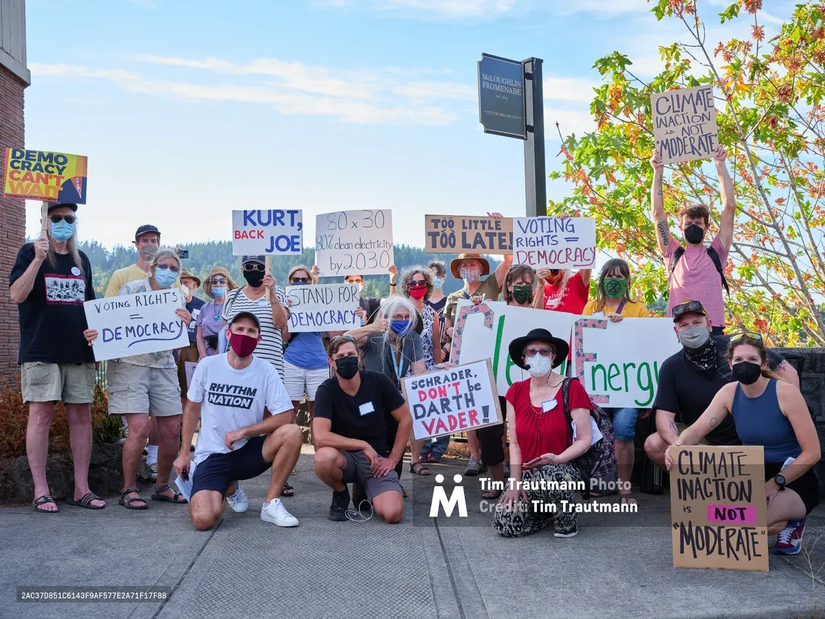 Masked protesters gather in a carefully arranged group portrait on the sidewalk outside Representative Kurt Schrader's Oregon City office, their handmade signs demanding climate action and voting rights creating a mosaic of grassroots activism. The late afternoon light bathes the diverse crowd of approximately twenty participants, ranging from young adults to seniors, all unified in their call for clean energy policy and democratic reform. Their colorful protest signs, featuring messages like "Climate Inaction is NOT Moderate" and "Kurt, Back Joe," stand out against the backdrop of brick buildings and autumn foliage along McLoughlin Promenade.