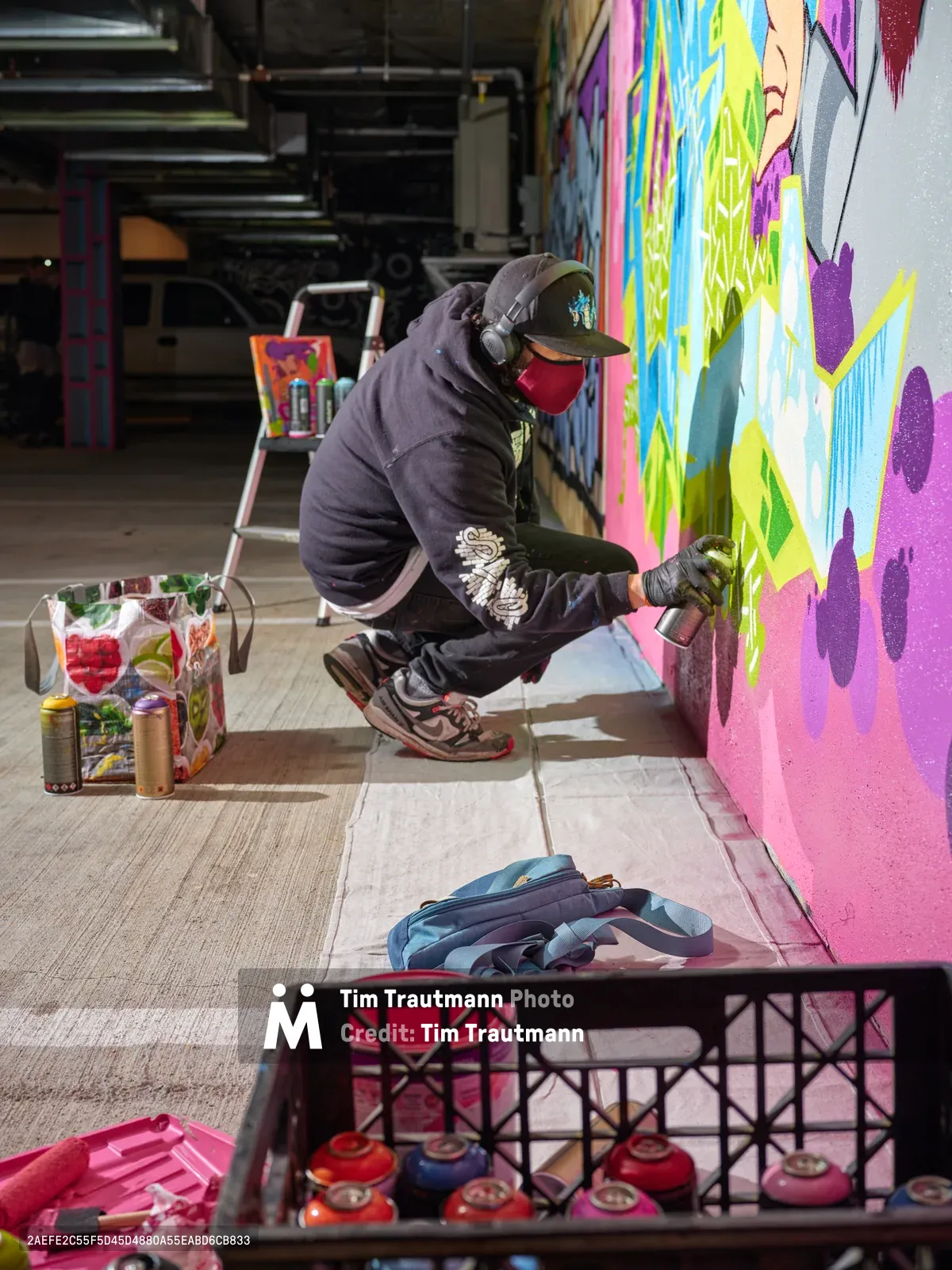 A masked street artist crouches intently beside a vibrant mural in progress, spray can in hand, within the industrial confines of Clay Creative's Southeast Portland parking garage. The scene captures the focused concentration of urban art creation, surrounded by crates of colorful spray paint and the raw concrete textures of the converted creative space. Brilliant neon pinks, electric blues, and lime greens cascade across the wall, contrasting sharply with the artist's dark hoodie and the garage's utilitarian atmosphere. The composition frames the intersection of industrial architecture and contemporary street art culture in Portland's creative district.