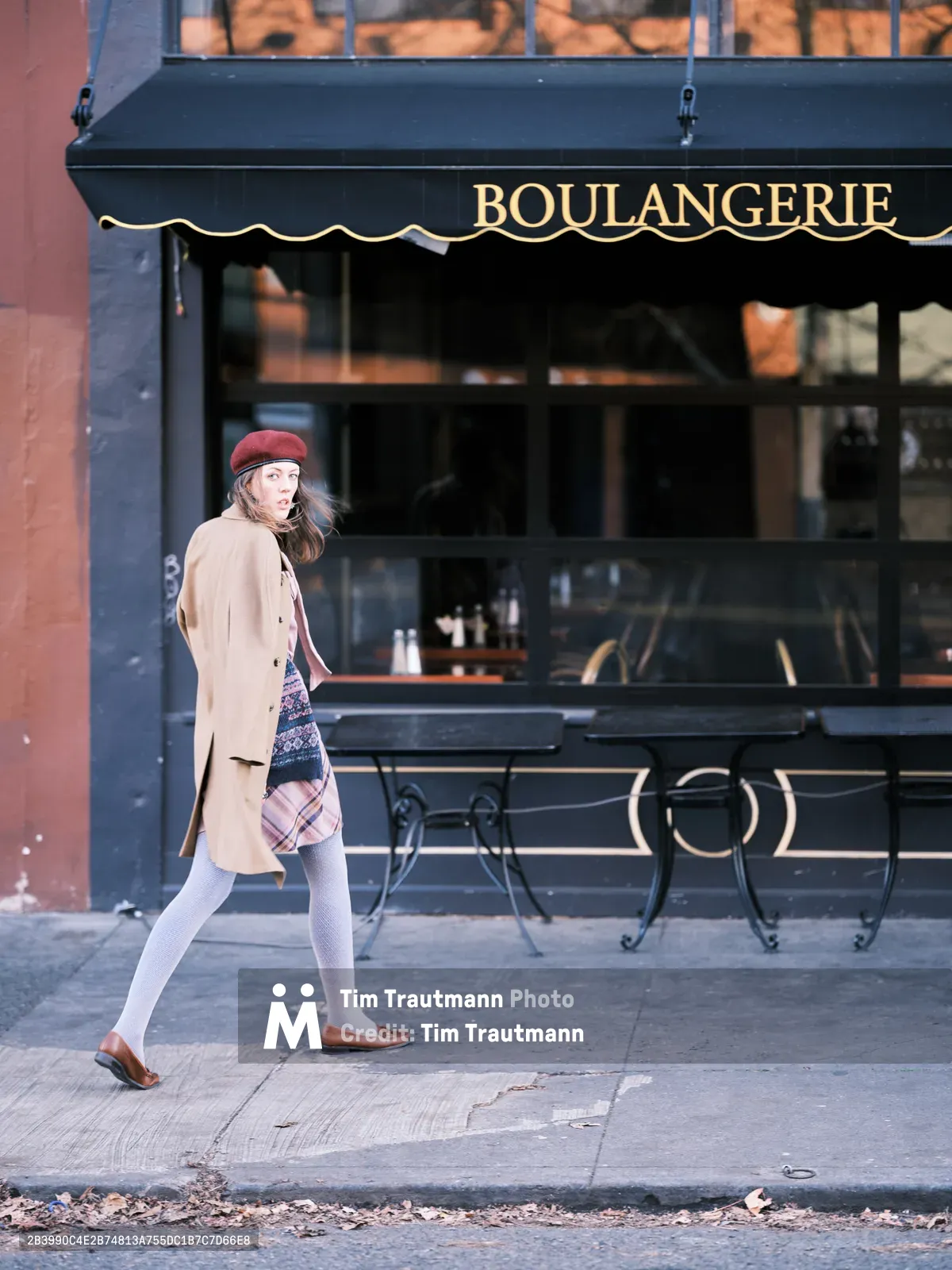 A young woman in a burgundy beret and camel coat strides past a French boulangerie on Northeast Alberta Street in Portland's Alberta district. Her outfit combines classic Parisian elements—the wool beret, tailored coat, and plaid skirt—with comfortable walking attire as she moves along the weathered sidewalk. The dark storefront with its elegant gold lettering creates an atmospheric backdrop, while natural light catches her mid-stride, suggesting a spontaneous street photography moment in this gentrified neighborhood.