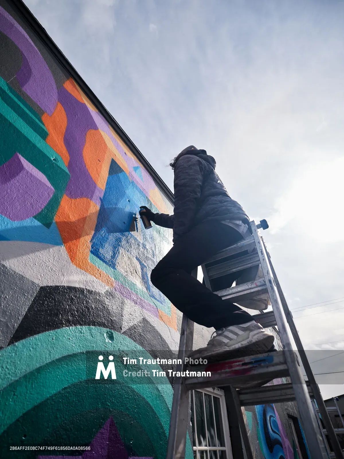 A bundled artist perches on aluminum scaffolding against a cloudy Pacific Northwest sky, spray painting an explosive kaleidoscope of geometric shapes across a building wall in Northeast Portland. The mural blooms with emerald greens, electric purples, burnt oranges, and azure blues, creating bold abstract forms that transform the urban landscape. The low-angle composition emphasizes the scale of the artistic endeavor while soft overcast light bathes the scene in muted tones typical of Portland's atmospheric conditions.