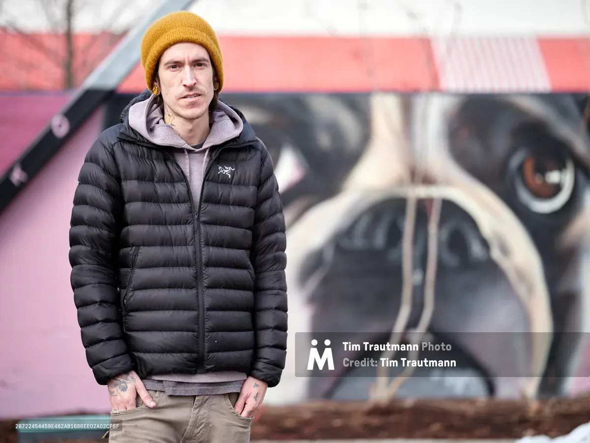 A portrait of muralist Kyle Danley, also known as Wetiko, standing in front of one of his large-scale murals depicting a close-up of a French Bulldog's face on a pink building. He wears a black Arc'teryx puffer jacket, a grey hoodie, khaki jeans, and a mustard yellow beanie. Neck and hand tattoos are visible. He looks directly into the camera with a calm, serious expression, hands in his pockets.