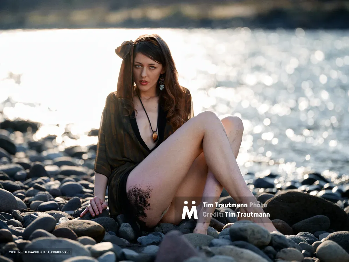 A young woman with flowing brunette hair sits contemplatively among smooth river stones along the Columbia River near Multnomah County's Floodplain Trail. Draped in a dark wrap that catches the warm backlight, she gazes pensively toward the sparkling water as golden hour light creates dramatic rim lighting around her silhouette. The rocky riverbed stretches into soft focus behind her, while dancing reflections on the water's surface complete this intimate riverside portrait.