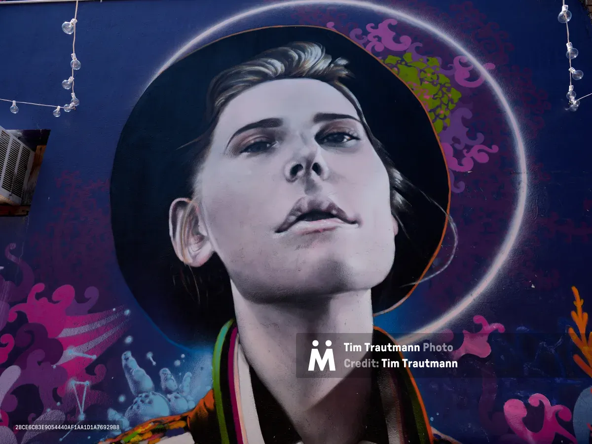 A striking street art portrait emerges from a cosmic backdrop in a Richmond neighborhood alley. The hyperrealistic face, rendered in monochromatic tones, gazes upward with serene contemplation while surrounded by vibrant purple and pink nebula-like formations. String lights delicately frame the composition against the deep blue wall, creating an intimate gallery atmosphere in this Southeast Division corridor.