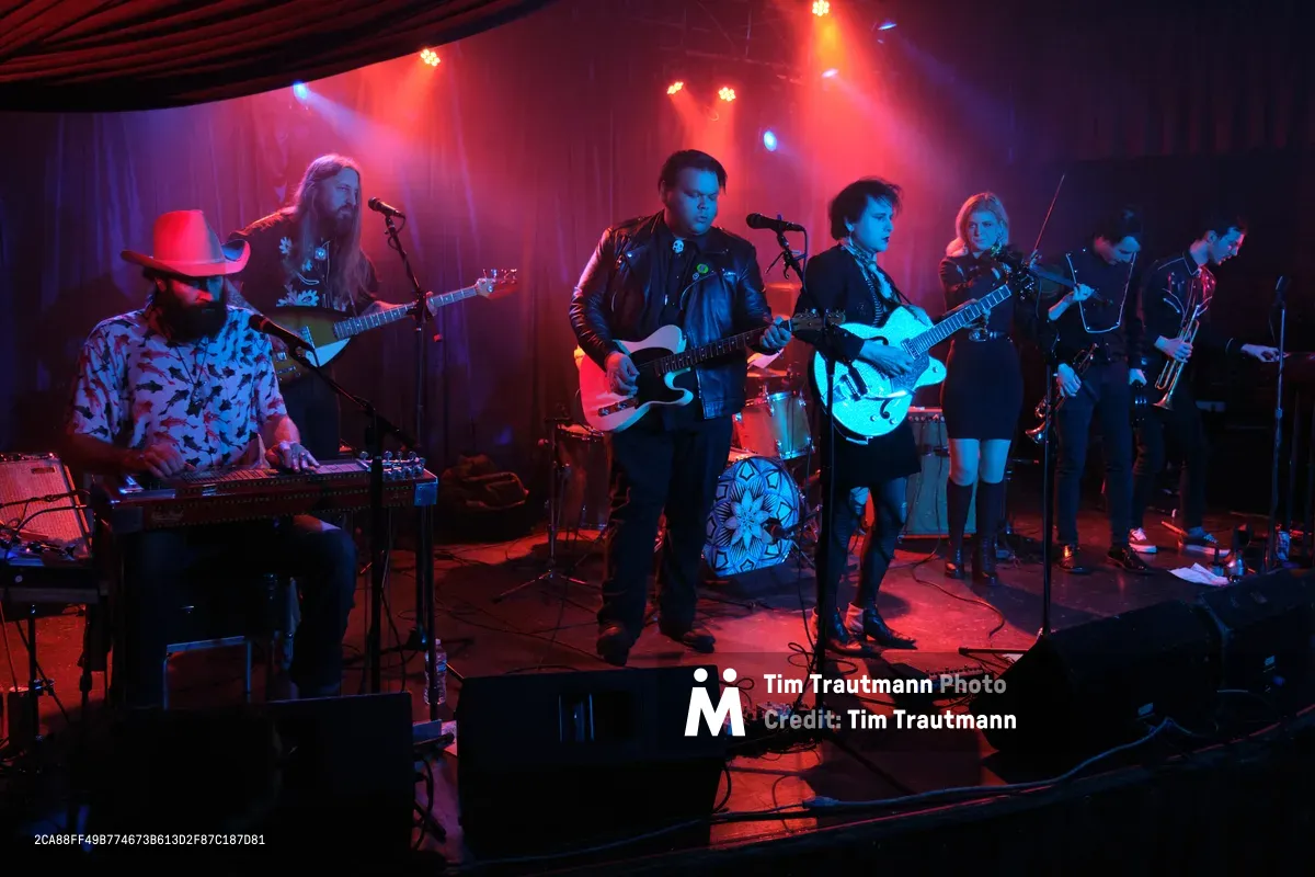 Bathed in crimson and azure stage lighting, six-piece band Roselit Bone delivers an intimate performance at the legendary Dante's venue in Portland, Oregon. The atmospheric lighting cuts through wisps of stage fog, illuminating the diverse ensemble of musicians wielding guitars, keyboards, and brass instruments. From left to right, a bearded keyboardist in a cowboy hat anchors the sound while electric guitars and horns create layers of melody across the small stage, their silhouettes dramatically carved by the moody concert illumination.