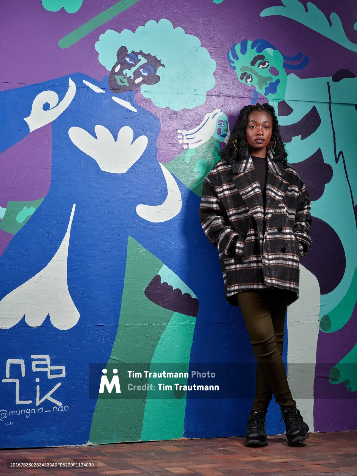 Naomi Likayi stands confidently before her vibrant street art mural on the boarded World Trade Center in downtown Portland. The young Black artist wears an oversized plaid coat against the cool Pacific Northwest air, her figure juxtaposed against swirling blues, purples, and teals of her abstract figurative work. The composition captures both the intimate scale of the artist and the bold public statement of her commissioned piece, embodying Portland's thriving street art culture.