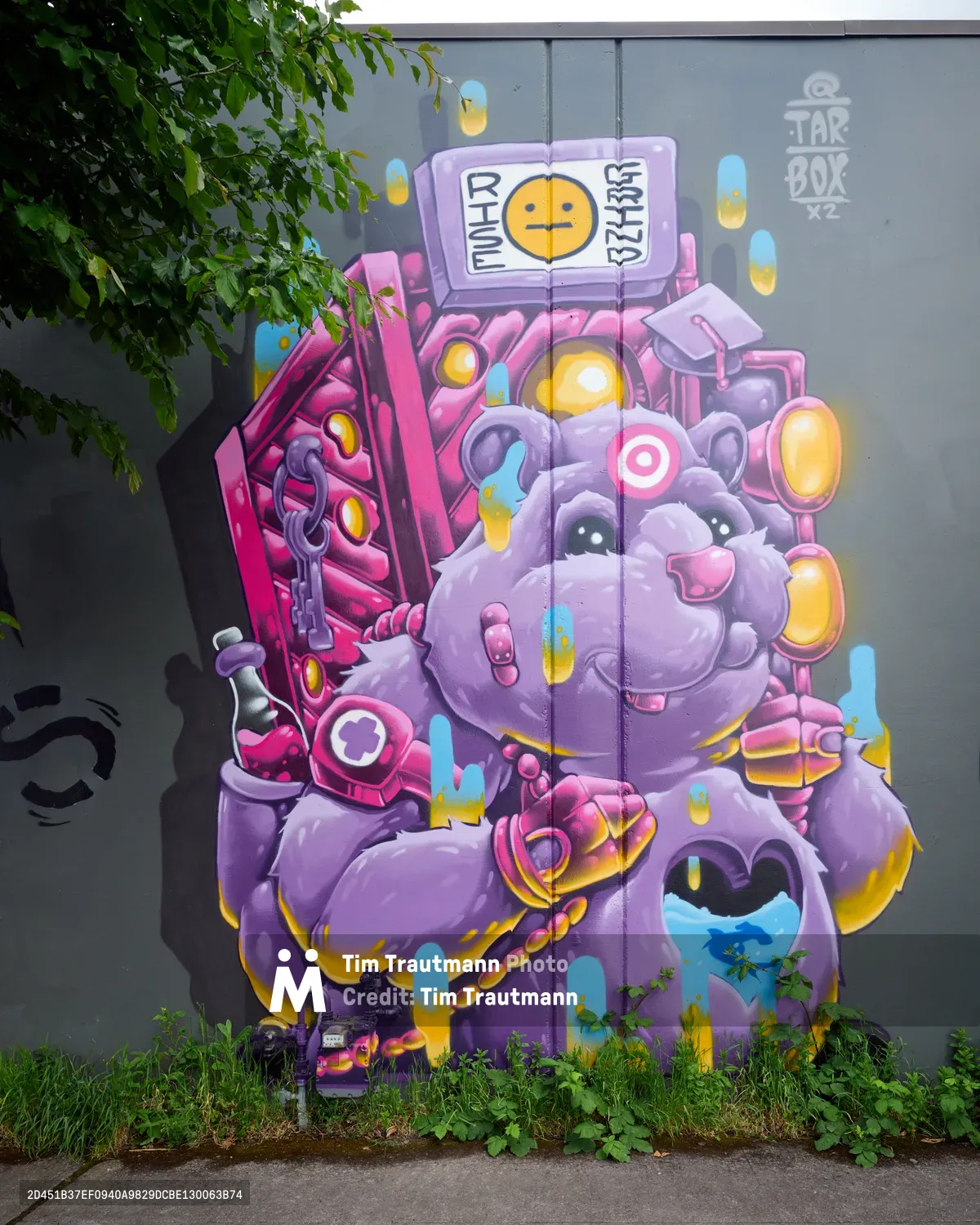A towering street art masterpiece dominates a concrete wall in Portland's Kerns neighborhood, where cyberpunk aesthetics meet whimsical character design. The mural depicts a purple robotic creature adorned with technological appendages, glowing screens, and neon-bright accents in magenta and yellow. Natural foliage frames the artwork, with overhanging tree branches creating an organic contrast against the digital fantasy below, while urban vegetation reclaims the base of the wall.