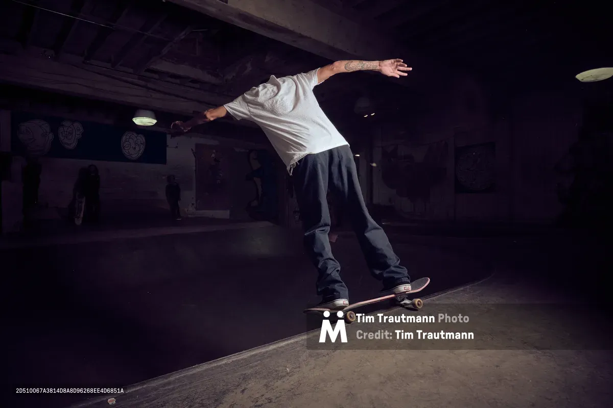 A tattooed skateboarder executes a fluid trick in the moody underground space of Commonwealth Skateboarding in Portland's Buckman neighborhood. The dimly lit basement venue creates dramatic shadows across weathered concrete, while exposed ceiling beams and vintage fixtures frame the athlete's precise movement. The skater's white shirt contrasts against the dark industrial interior, capturing the raw authenticity of Portland's skateboarding culture.