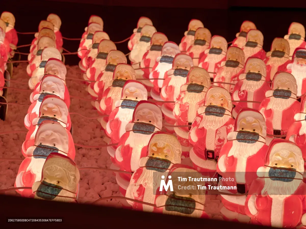 Rows of identical plastic Santa Claus figures stretch across a display in Portland's Sellwood-Moreland neighborhood, each wearing a black face mask in adaptation to COVID-19 protocols. The warm red glow of holiday lighting bathes the mass-produced Christmas decorations, creating an eerie yet poignant commentary on how even beloved seasonal traditions were transformed by the pandemic. The repetitive pattern of masked Santas extends into the background, suggesting both holiday commerce and collective adaptation to unprecedented times.