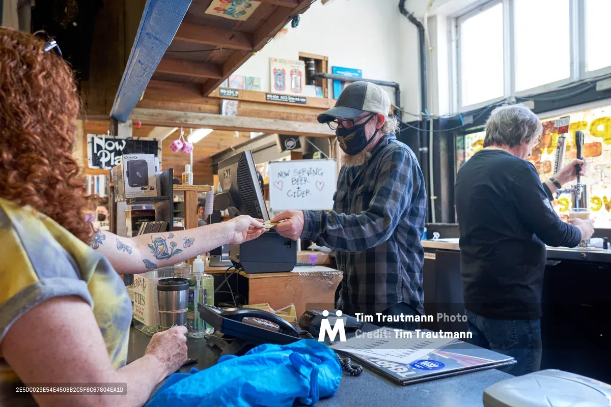 Inside the warm, rustic interior of Music Millennium on East Burnside during Record Store Day 2022, a masked clerk in a gray trucker cap and plaid flannel shirt processes a transaction with a tattooed customer. Exposed wooden beams frame the scene while vinyl records and merchandise create an authentic record store atmosphere. Natural light streams through windows, illuminating this intimate moment of independent retail commerce in Portland's Kerns neighborhood.