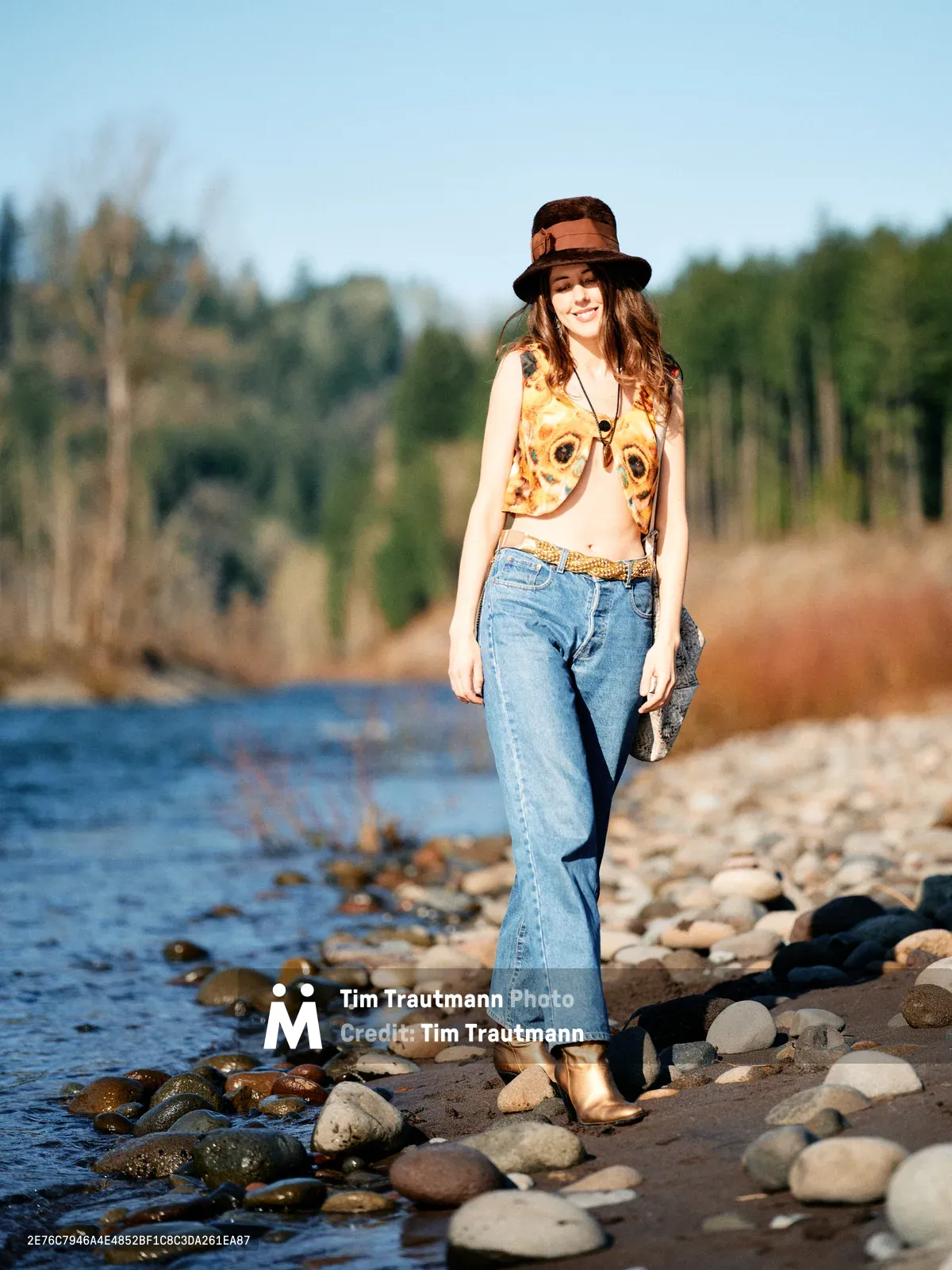 A young woman with flowing brown hair stands confidently on the rocky shoreline of Oregon's Floodplain Trail, her vintage-inspired outfit creating striking contrast against the natural landscape. Golden hour light illuminates her brown fedora hat, graphic vest, and high-waisted jeans as she poses beside the gentle waters. The composition captures her relaxed stance among smooth river stones, with the soft-focus forest backdrop emphasizing the connection between fashion and nature. Her warm smile and bohemian styling evoke a sense of free-spirited adventure in this serene riparian setting.
