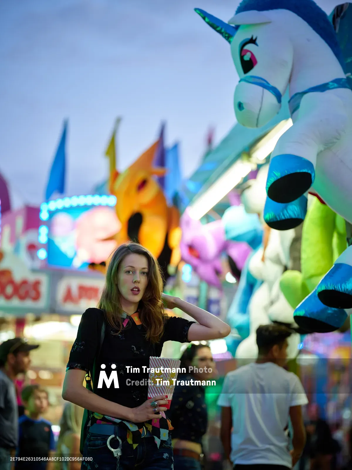 A young woman with flowing brown hair stands contemplatively in the heart of Clark County Fair in Vancouver, Washington, clutching a striped popcorn container as neon carnival lights paint the dusky sky behind her. Towering inflatable attractions, including a whimsical unicorn bouncer, create a dreamlike backdrop of carnival magic. The shallow depth of field isolates her pensive expression against the bustling fairground atmosphere, where other visitors blur into the enchanting chaos of evening entertainment.