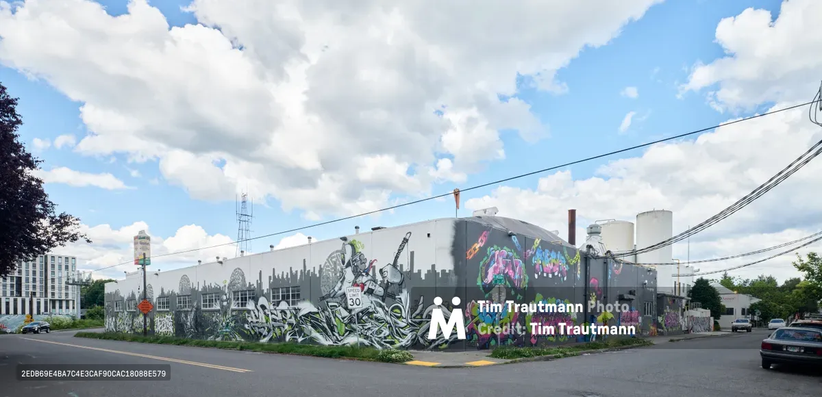 The sprawling concrete facade of Portland's former Sunshine Dairy building transforms into a vibrant street art gallery in the Kerns neighborhood. Elaborate murals cascade across the industrial structure, featuring bold graffiti lettering, cartoon-like characters in electric blues and magentas, and a striking cityscape silhouette that mirrors the urban environment. Power lines slice through partly cloudy skies above this corner intersection on Northeast 21st Avenue, where contemporary street culture breathes new life into industrial heritage.