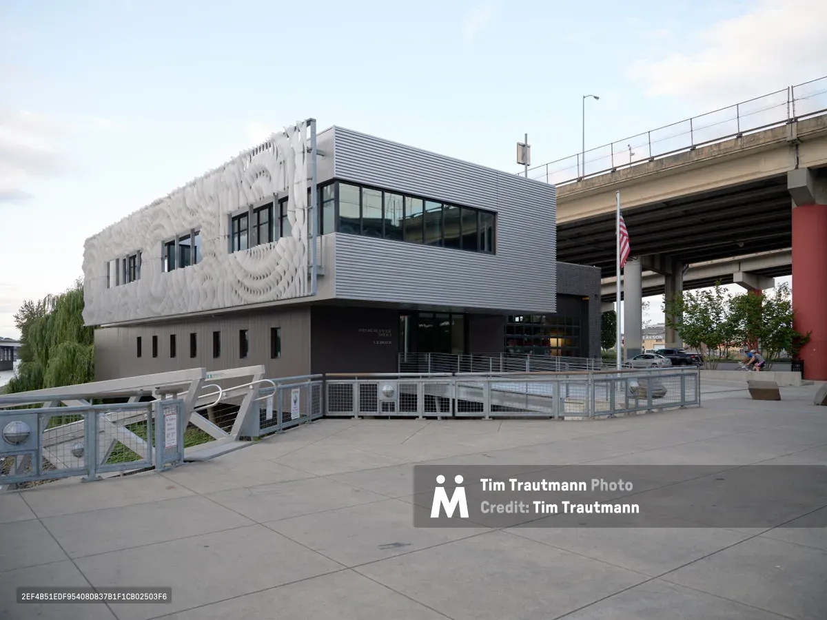 Portland Fire & Rescue Station 21 emerges as a striking example of contemporary public architecture beneath the Interstate 84 overpass in the Central Eastside district. The building's sculptural white facade features an undulating parametric pattern that contrasts dramatically with the corrugated metal upper level and industrial concrete surroundings. Captured during the golden hour transition, the structure's modern design philosophy transforms utilitarian emergency services architecture into an urban landmark that bridges infrastructure and community.