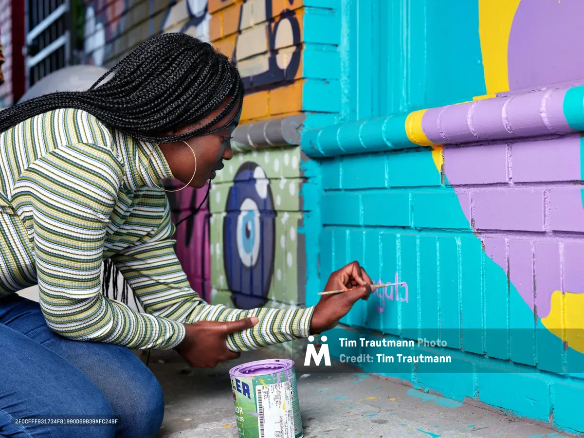 Naomi Likayi kneels beside her vibrant street mural in Portland's Alberta Arts District, carefully painting her signature in purple letters on a brilliant turquoise section of the wall. Her intricate braided hair catches the afternoon light as she concentrates on her work, wearing a striped long-sleeve shirt and jeans. Behind her, the colorful geometric mural explodes across the brick wall in bold strokes of purple, yellow, and cyan, transforming the urban corner of Northeast 15th and Killingsworth into a canvas of community expression.